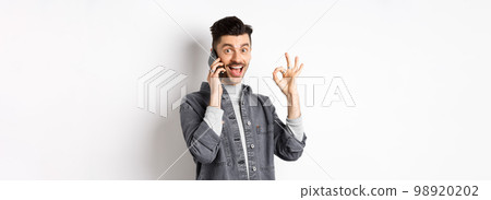 Excited young man talking on phone and showing okay gesture, smiling satisfied, standing against white background 98920202