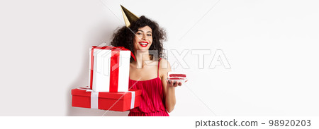 Happy birthday girl in red dress, celebrating and holding gifts with bday cake, standing on white background 98920203