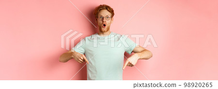 Image of amazed young man with red hair and beard, wearing glasses and t-shirt, pointing fingers down and staring excited at camera, pink background 98920265