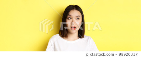 Close up of beautiful asian woman with clean perfect skin, raising eyebrow and looking left with intrigued face, standing over yellow background Close up of beautiful asian woman with clean perfect skin, raising eyebrow and looking left with intrigued face, standing over yellow background 98920317