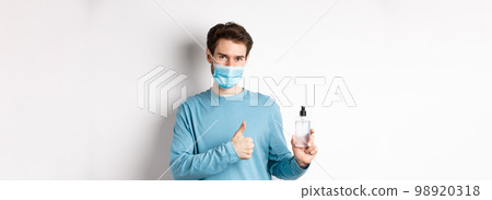 Covid-19, health and quarantine concept. Handsome smiling man in face mask, showing thumbs up and hand sanitizer bottle, recommending antiseptic brand, white background Covid-19, health and quarantine concept. Handsome smiling man in face mask, showing thumbs up and hand sanitizer bottle, recommending antiseptic brand, white background 98920318
