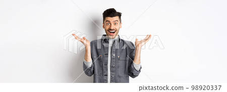 Excited young man cheering from good news, spread hands sideways and looking happy, smiling amazed, standing on white background Excited young man cheering from good news, spread hands sideways and looking happy, smiling amazed, standing on white background 98920337