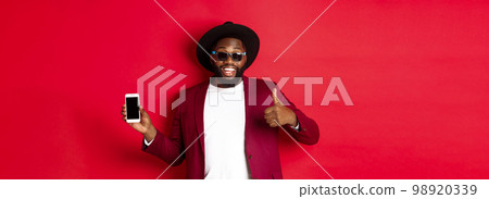 Handsome and stylish Black man showing phone screen and thumb up at camera, recommending online store app, red background 98920339