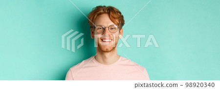 Close up of handsome redhead man in glasses looking at camera, smiling with white teeth, standing against mint background Close up of handsome redhead man in glasses looking at camera, smiling with white teeth, standing against mint background 98920340