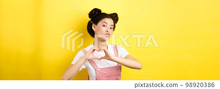 Silly japanese girl showing heart gesture and wishing happy Valentines day, pucker lips to kiss lover, standing on yellow background 98920386