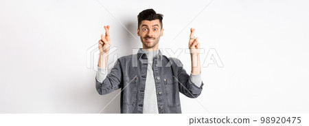 Hope dream come true. Positive and handsome guy making wish with fingers crossed, looking hopeful and smiling at camera, waiting results, standing on white background 98920475