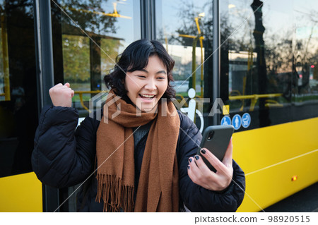 Portrait of cheerful asian girl talks on mobile phone, video chats, looks amazed at smartphone camera, stands on bus stop Portrait of cheerful asian girl talks on mobile phone, video chats, looks amazed at smartphone camera, stands on bus stop 98920515