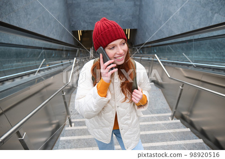 Portrait of young redhead woman walks around city, goes up stairs with mobile phone, talks on smartphone and smiles 98920516