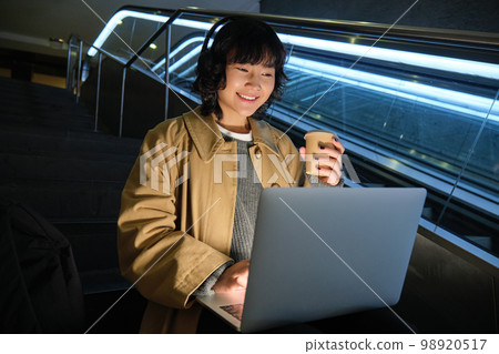 Young hipster girl in headphones, sits on staircase, drinks coffee and works on her laptop, studies in public place 98920517