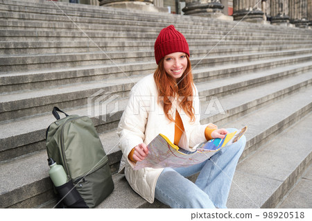 Smiling girl traveler, tourist sits on stairs near museum and holds paper map, explores city during sightseeing adventure trip Smiling girl traveler, tourist sits on stairs near museum and holds paper map, explores city during sightseeing adventure trip 98920518