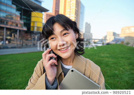 University lifestyle. Young asian student, girl with tablet and notebook, talks on mobile phone while walks from campus on street University lifestyle. Young asian student, girl with tablet and notebook, talks on mobile phone while walks from campus on street 98920521