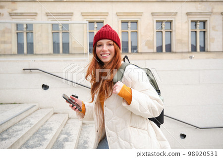Smiling redhead girl, student tourist walks around city, goes up the stairs, looks at mobile phone map to get around town, sends message on smartphone 98920531