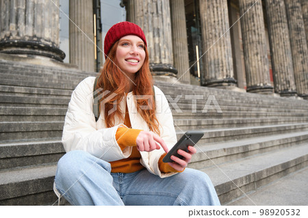Portrait of young urban girl in red hat, sits on stairs near museum, holds mobile phone, connects to public wifi and surfs net, uses smartphone apps 98920532