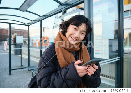 Cute smiling asian girl standing on bus stop, holding smartphone, wearing winter jacket and scarf. Woman commuting to work or university via public transport, stands on road 98920534