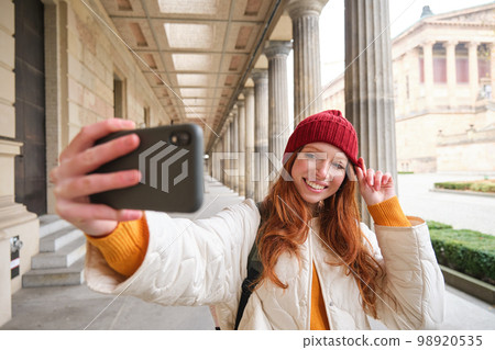 Stylish redhead girl tourist, takes selfie in front of tourism attraction, makes photo with smartphone, looks at mobile camera and poses 98920535