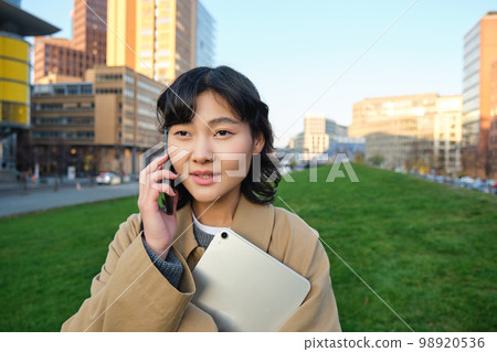 Stylish smiling girl, university student in trench coat, holds tablet, talks on mobile phone, has conversation over telephone and looks relaxed, stands on street 98920536