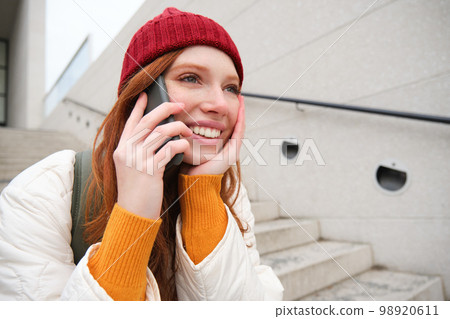 Beautiful smiling redhead female model, sits on street and talks on mobile phone, uses smartphone app to call abroad, laughing during telephone conversation 98920611