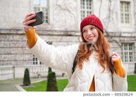 Cute ginger girl in red hat takes selfie during her tourist trip abroad. Young redhead woman makes a photo of herself in front of historical landmark 98920624