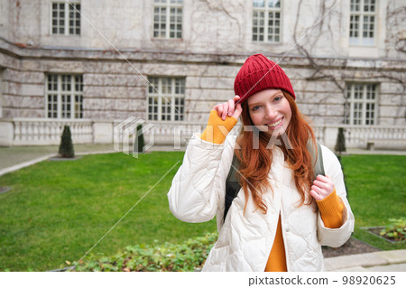 Beautiful redhead woman with backpack, tourist walking around city park and looks around, wears red hat and warm coat 98920625