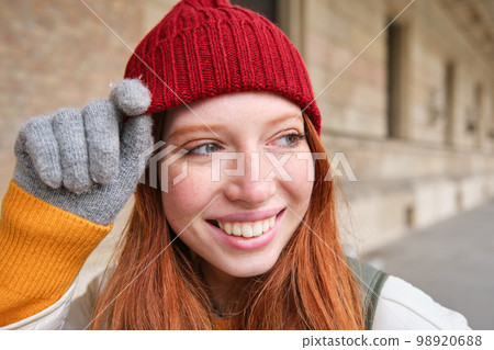 Headshot of happy redhead girl with freckles, wears red hat and gloves in winter, walks around city on chilly weather and smiles 98920688