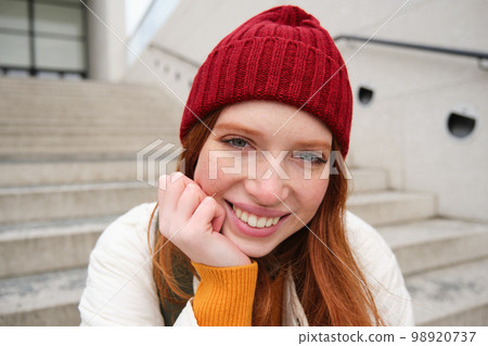 Beautiful redhead student, girl in red hat, smiles sincere, looks happy and relaxed, sits on stairs outdoors 98920737