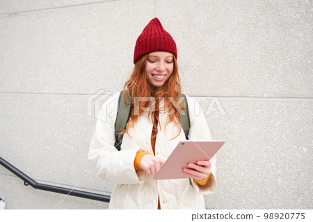 Young redhead woman with red hat, uses her digital tablet outdoors, stands on street with gadget, connects to wifi internet and searches for a location in internet Young redhead woman with red hat, uses her digital tablet outdoors, stands on street with gadget, connects to wifi internet and searches for a location in internet 98920775