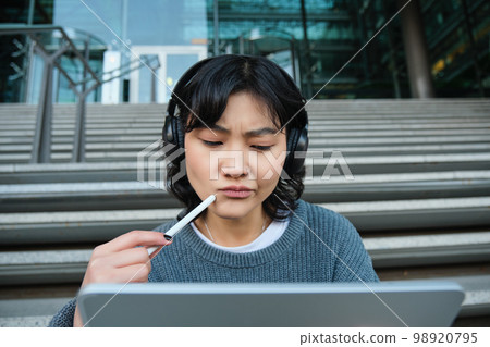Portrait of asian girl with troubled face, draws on digital tablet with pen, looks complicated at her work, feels stuck no inspiration, sits on stairs and thinks Portrait of asian girl with troubled face, draws on digital tablet with pen, looks complicated at her work, feels stuck no inspiration, sits on stairs and thinks 98920795