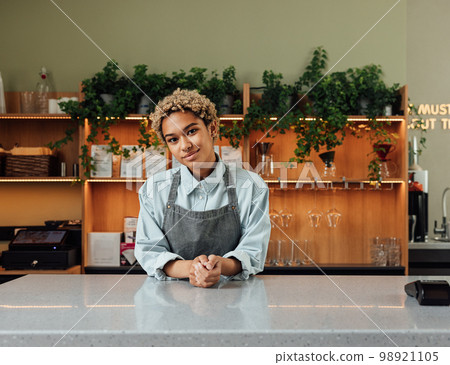 Barista in an apron with short curly hair leaning on a counter. Coffeeshop owner looking at camera. Barista in an apron with short curly hair leaning on a counter. Coffeeshop owner looking at camera. 98921105