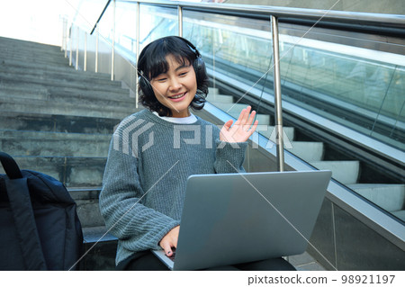 Smiling young student, wears headphones, sits on street stairs and waves hand at her laptop, connects to video chat, conference or training session on remote Smiling young student, wears headphones, sits on street stairs and waves hand at her laptop, connects to video chat, conference or training session on remote 98921197