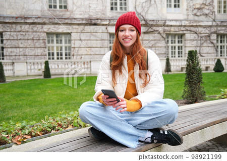 Portrait of stylish young woman, 25 years, sits on bench in park and uses mobile phone, reads online news, messages or watches video on smartphone app, connects to public wifi 98921199
