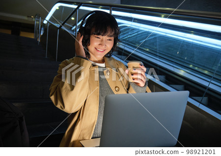 Beautiful young girl student, korean woman sits on staircase, watches video on laptop in headphones, drinks takeaway coffee, works in public place on remote Beautiful young girl student, korean woman sits on staircase, watches video on laptop in headphones, drinks takeaway coffee, works in public place on remote 98921201