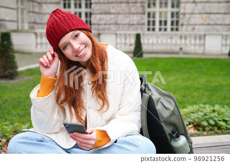 Young smiling redhead girl sits in park with smartphone, texting message on mobile phone, using telephone, connecting to public wifi and surfing internet 98921256