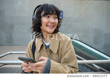 Technology and people. Young asian girl, student standing on street in headphones, listens music and smiling, chats on mobile phone, sends a text message, waits for someone on street Technology and people. Young asian girl, student standing on street in headphones, listens music and smiling, chats on mobile phone, sends a text message, waits for someone on street 98921377