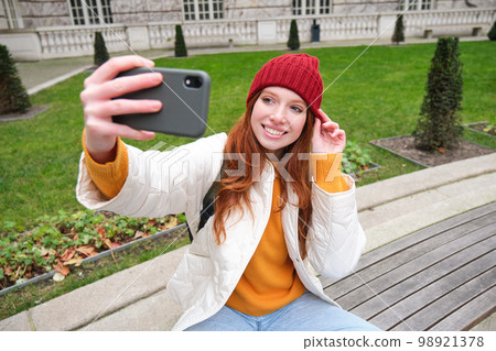 Young teen redhead girl sits on bench in park and takes selfie, makes a photo of herself with smartphone app, records vlog 98921378