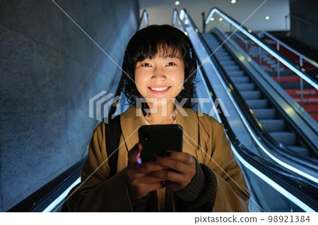 Portrait of girl student in headphones, listens music, commutes, goes down escalator, looks at smartphone with pleased smile, happy face 98921384
