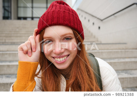 Stylish redhead girl in warm red hat, smiling relaxed, sitting with backpack on stairs near building, waits for someone outdoors Stylish redhead girl in warm red hat, smiling relaxed, sitting with backpack on stairs near building, waits for someone outdoors 98921388