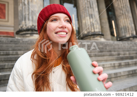 Portrait of redhead woman drinking from thermos, sitting on street stairs and enjoys hot drink from flask 98921412