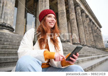 Portrait of young urban girl in red hat, sits on stairs near museum, holds mobile phone, connects to public wifi and surfs net, uses smartphone apps Portrait of young urban girl in red hat, sits on stairs near museum, holds mobile phone, connects to public wifi and surfs net, uses smartphone apps 98921419