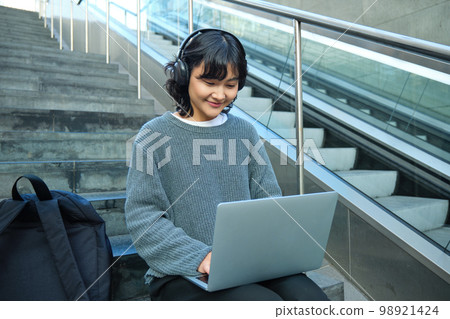 Portrait of beautiful young korean woman, student sits on stairs in public place, listens music in headphones, works on project on laptop, works remotely Portrait of beautiful young korean woman, student sits on stairs in public place, listens music in headphones, works on project on laptop, works remotely 98921424