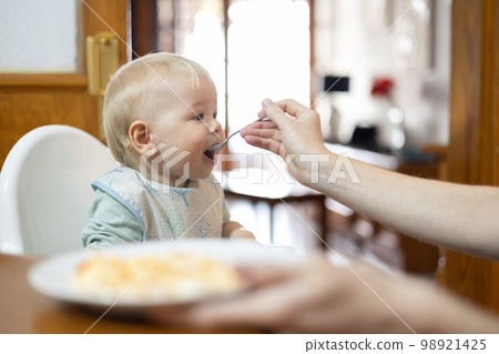 Mother spoon feeding her infant baby boy child sitting in high chair at the dining table in kitchen at home. Mother spoon feeding her infant baby boy child sitting in high chair at the dining table in kitchen at home. 98921425