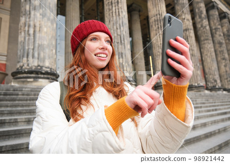 Portrait of young blogger, redhead girl tourist takes pictures of sightseeing, makes photos on smartphone camera, records video and smiles Portrait of young blogger, redhead girl tourist takes pictures of sightseeing, makes photos on smartphone camera, records video and smiles 98921442