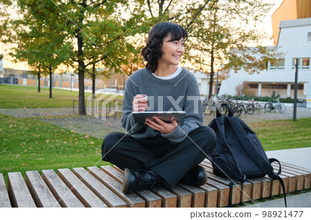 Young asian girl, student sits on bench, graphic designer with pencil and digital tablet draws scatches, does her homework outdoors Young asian girl, student sits on bench, graphic designer with pencil and digital tablet draws scatches, does her homework outdoors 98921477
