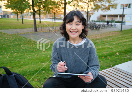 Young girl, korean artist or art student sits in park with digital tablet, draws with graphic pen, scatches a design or project, looks around Young girl, korean artist or art student sits in park with digital tablet, draws with graphic pen, scatches a design or project, looks around 98921480