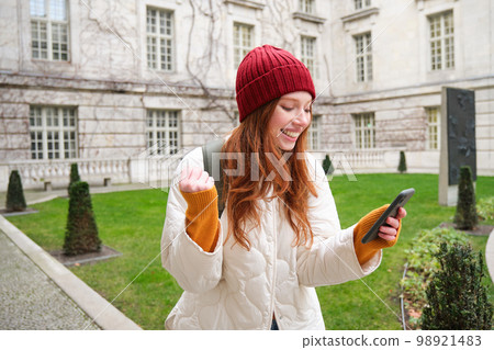 Portrait of cheerful redhead woman with smartphone, celebrating, reading amazing news and rejoicing, triumphing on street 98921483