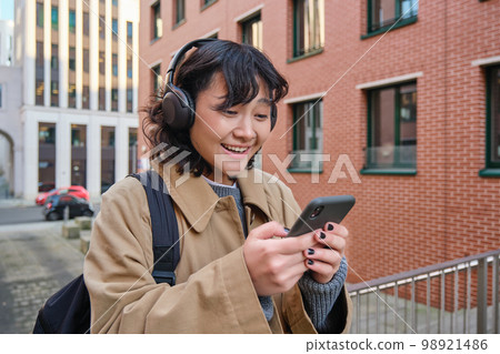 Happy korean girl looks at mobile phone and listens music in headphones, stands on street in city centre, reads text message on smartphone Happy korean girl looks at mobile phone and listens music in headphones, stands on street in city centre, reads text message on smartphone 98921486