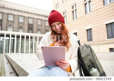 Redhead girl smiles, sits outdoors near building with digital tablet, thermos and backpack, connects to public internet and searches smth online on her gadget 98921549