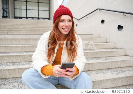 Redhead girl in red hat, sits on stairs and uses mobile phone. Modern woman holding smartphone, texting message, using telephone application outdoors 98921550
