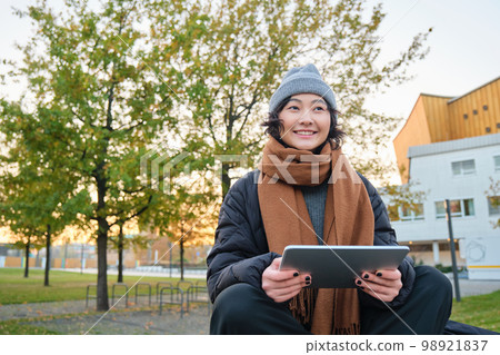 Portrait of asian girl in warm clothes, sits on bench with digital tablet and graphic pen, smiling happily, draws outdoors in chilly weather 98921837