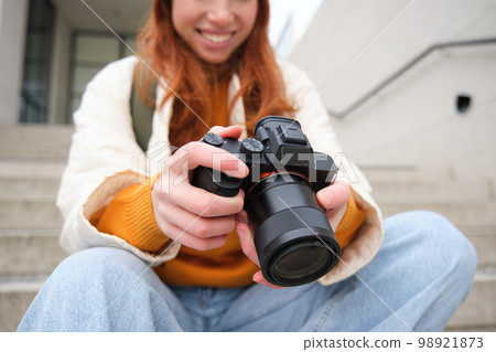 Smiling redhead girl photographer, checks her shots, holds camera and looks at screen, takes photos outdoors, walks around street and does streetstyle shooting Smiling redhead girl photographer, checks her shots, holds camera and looks at screen, takes photos outdoors, walks around street and does streetstyle shooting 98921873