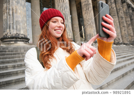 Portrait of young blogger, redhead girl tourist takes pictures of sightseeing, makes photos on smartphone camera, records video and smiles Portrait of young blogger, redhead girl tourist takes pictures of sightseeing, makes photos on smartphone camera, records video and smiles 98921891